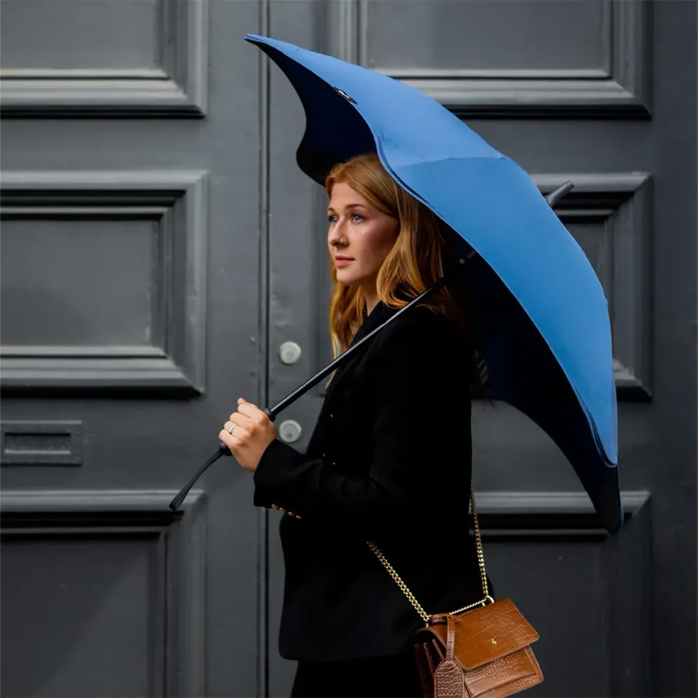 Woman holding BLUNT Coupe umbrella in navy while walking outdoors against dark grey doorway background