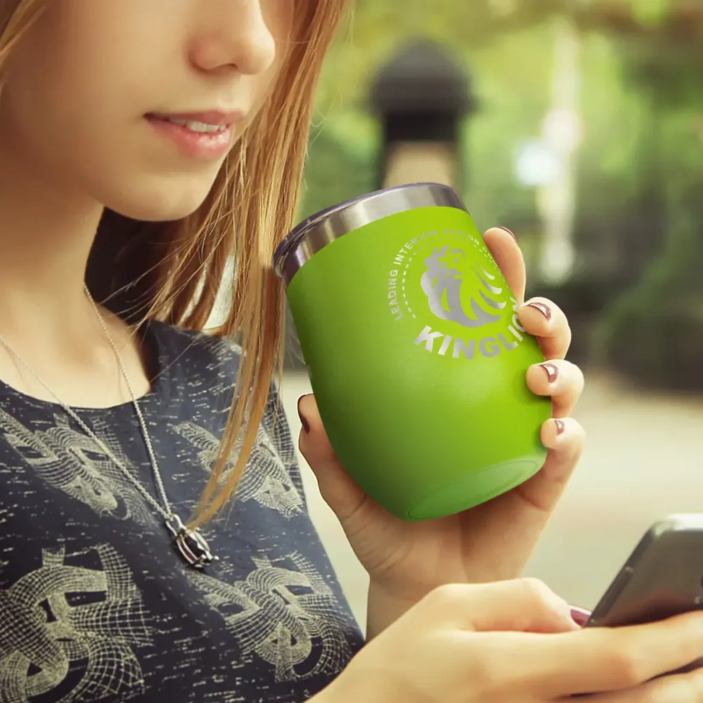 Woman holding green Cordia Vacuum Cup with printed logo while using smartphone outdoors.