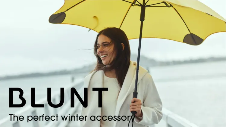 Smiling woman holding a yellow BLUNT umbrella on a windy pier, wearing a light coat