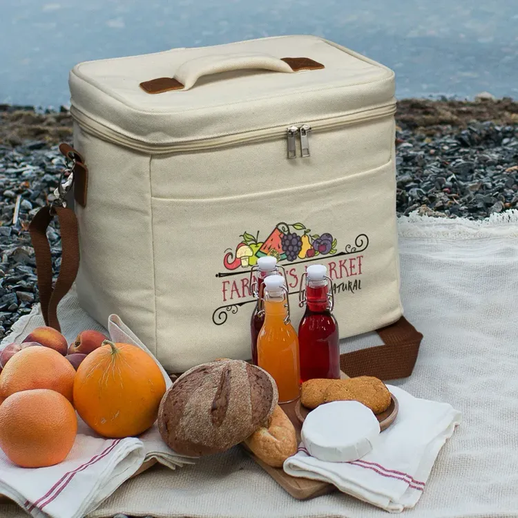 Colton Cooler Bag in natural canvas displayed outdoors with picnic food and drinks beside it.
