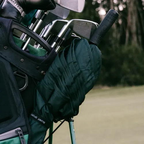Folded forest green BLUNT Sport umbrella placed in golf bag beside silver clubs outdoors