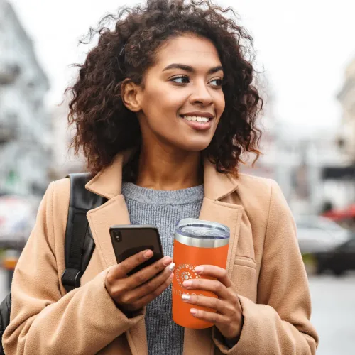 Woman outdoors holding reusable orange coffee cup with printed logo and smartphone.