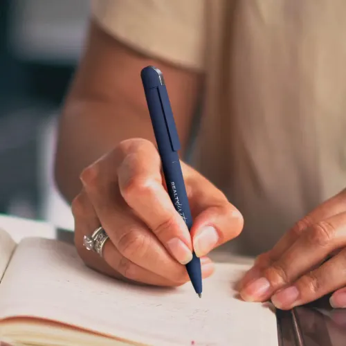 Person writing in notebook with navy metal pen engraved with logo.