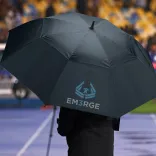 Person holding navy Adventura Sports umbrella with branded panel at outdoor sports venue.