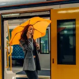 Woman standing by train door holding mango orange BLUNT Metro umbrella