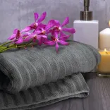 Folded charcoal Palms Luxury Towels with orchid flowers, candle, and dispenser on wooden surface.